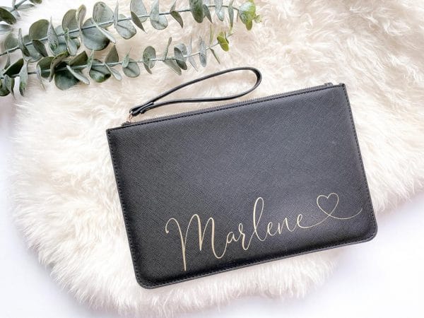 Schwarze Clutch liegt auf einem weißen Schaffell. Sie ist personalisiert mit einem goldenen Aufdruck "Marlene"